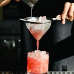 bartender pouring a pink cocktail out of a shaker, through a strainer, and into a rocks glass