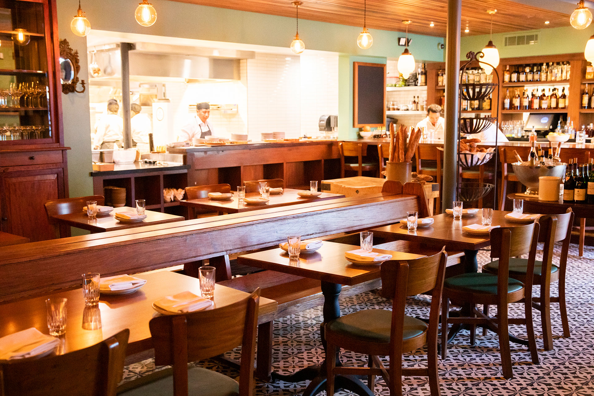 bistro-dining-room-scaled shot at an angle of the bicyclette dining room, with wooden chairs, tables, and benches arranged in a row in front of an open kitchen and an open bar with staff prepping their various stations