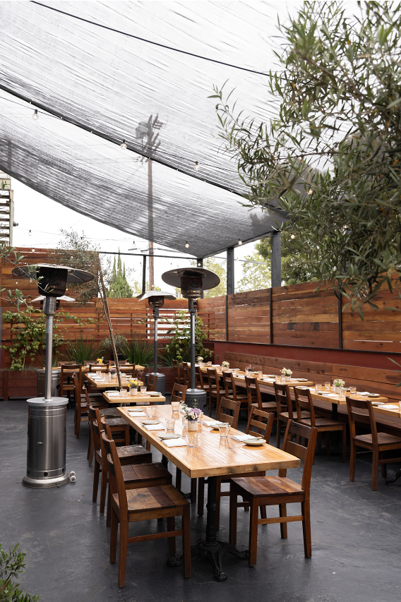 Outdoor patio with shade canopy, olive trees, heat lamps, and wooden tables set for al fresco dining