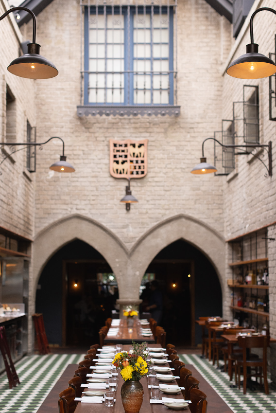 Long wooden table set for private dining beneath gothic arches with industrial pendant lights and yellow flower centerpiece