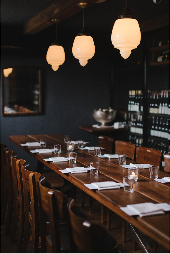 Intimate private dining room with dark walls, glowing pendant lights, and wooden table set with glassware and napkins