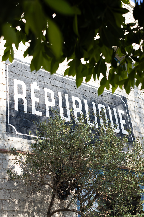 Exterior view of République sign painted on white brick wall framed by tree branches